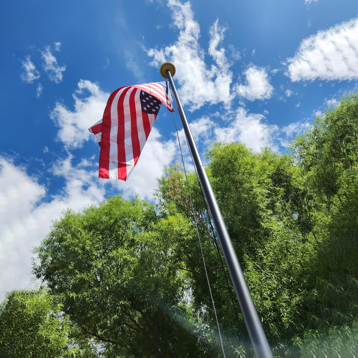 American flag on a pole against a blue sky with clouds and green trees.