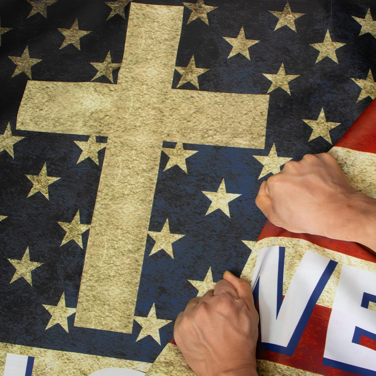A close-up of a patriotic flag or banner featuring a large gold cross on a dark blue field with gold stars, partially overlaid by hands holding the edges. The design also includes red and white sections with cutout letters.