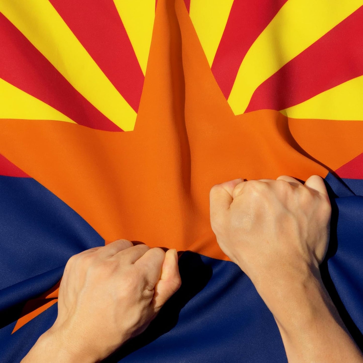 Close-up of a person's hands pulling apart an Arizona state flag, showing the red and yellow rays, orange sun, and blue lower section.